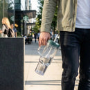 Person holding a clear water bottle with a FlavaFil with a green lid on a city street.