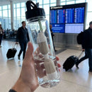 Clear water bottle with black cap held in a hand at an airport terminal with FlavaFil in, offering peace of mind as you travel and don't trust the water